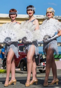 Three women in flapper dresses and feather fans pose next to a vintage car.