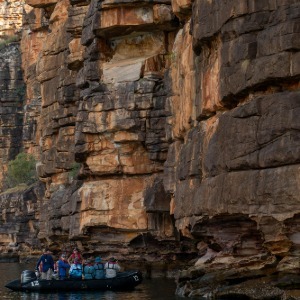 A small boat navigates towering, rugged red rock cliffs.