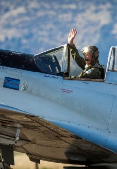 A pilot waves from the cockpit of a parked vintage propeller plane on a sunny day.