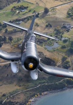 A grey warbird with a yellow nose flies low over a lake and green fields below.