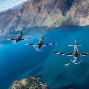 Three military planes fly in formation over a large, beautiful lake surrounded by mountains.