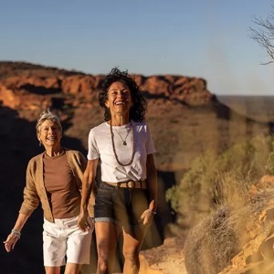 Two women smile as they hike a trail with a rock formation in the background.