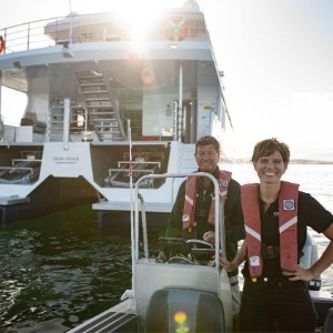 Two crew members steer a small boat with a larger vessel behind.