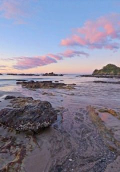 A beach is covered in rocks and tidal pools at sunset under a pink and blue sky.