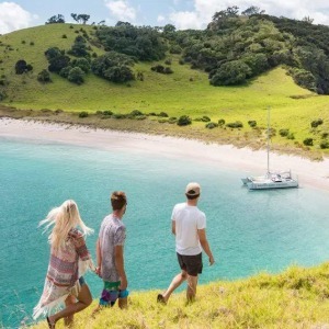 Three people descend a grassy hill toward a secluded cove with a catamaran.