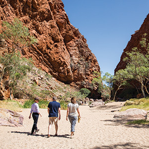 Three people walking through a sandy, narrow red-rock gorge.