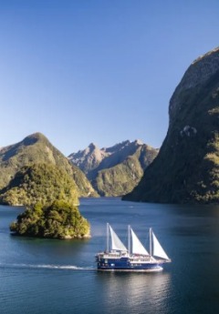 A boat with three white sails cruises past a small island between steep fjords.