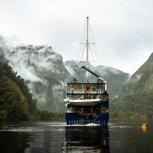 The stern of a large blue boat faces away, cruising through a misty fjord valley.