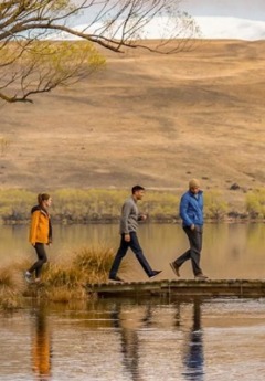 Three people walk on a wooden walkway over a small pond with hills in the background.