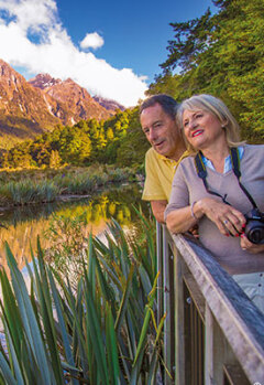 An older couple, with a camera, stands on a boardwalk admiring the mountains.