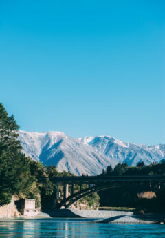 Three people stand distantly on a bridge stretching over blue water. There are snowy capped mountains in the background