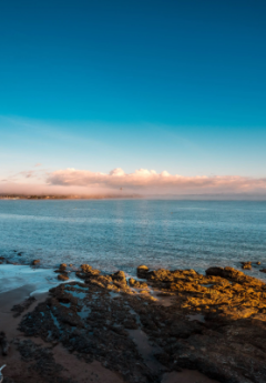 Sunrise illuminates rocky beach, calm sea, and distant clouds