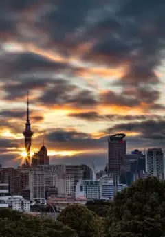 The sun sets behind the Sky Tower in Auckland, lighting up the cloudy city skyline in a golden hue