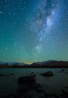 The nebulous clouds of the Milky Way shine over Mt Cook National Park, with snowcapped mountains in the background, and a pale turquoise lake in the foreground.