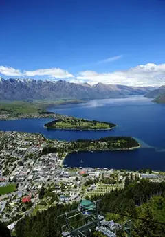 A bird's eye view of Queenstown from Skyline, with a blue sky dotted with fluffy white clouds, large snowcapped mountains, a deep blue lake, and the city along the lakeshore.