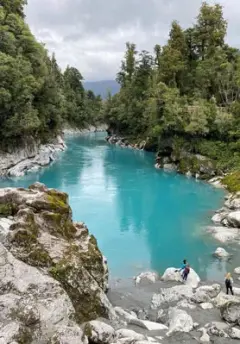 A bright blue lake disapears down a gorge and people climb on the silver rocks surrounding the lake