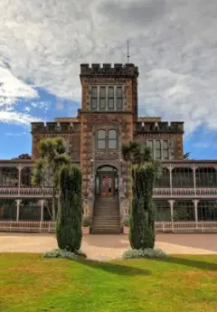 Larnach Castle under cloudy blue skies with two tall bushes standing at either side of the entrance