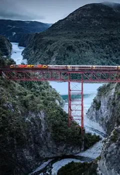 TranzAlpine Train crossing a river, surrounded by stunning natural scenery.